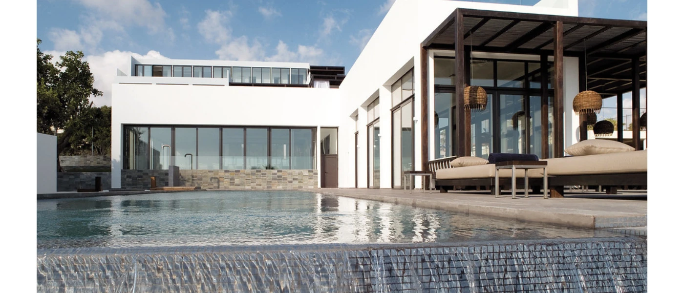 Infinity pool edge next to cushioned loungers overlooked by a building with full-length windows