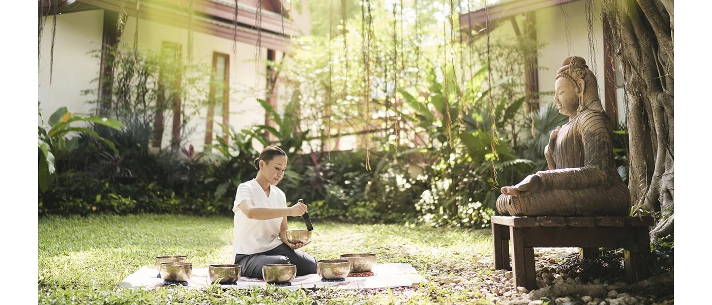 A woman sits on a blanket in a tropical garden, using gold sound healing bowls in front of a Buddha statue