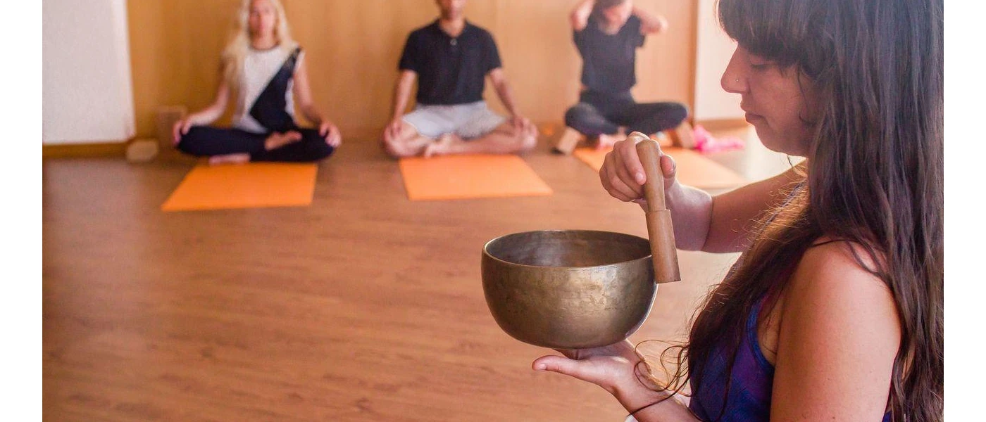 Three people sit cross legged on orange mats in an airy studio, as a woman at the front of the class play a sound bowl