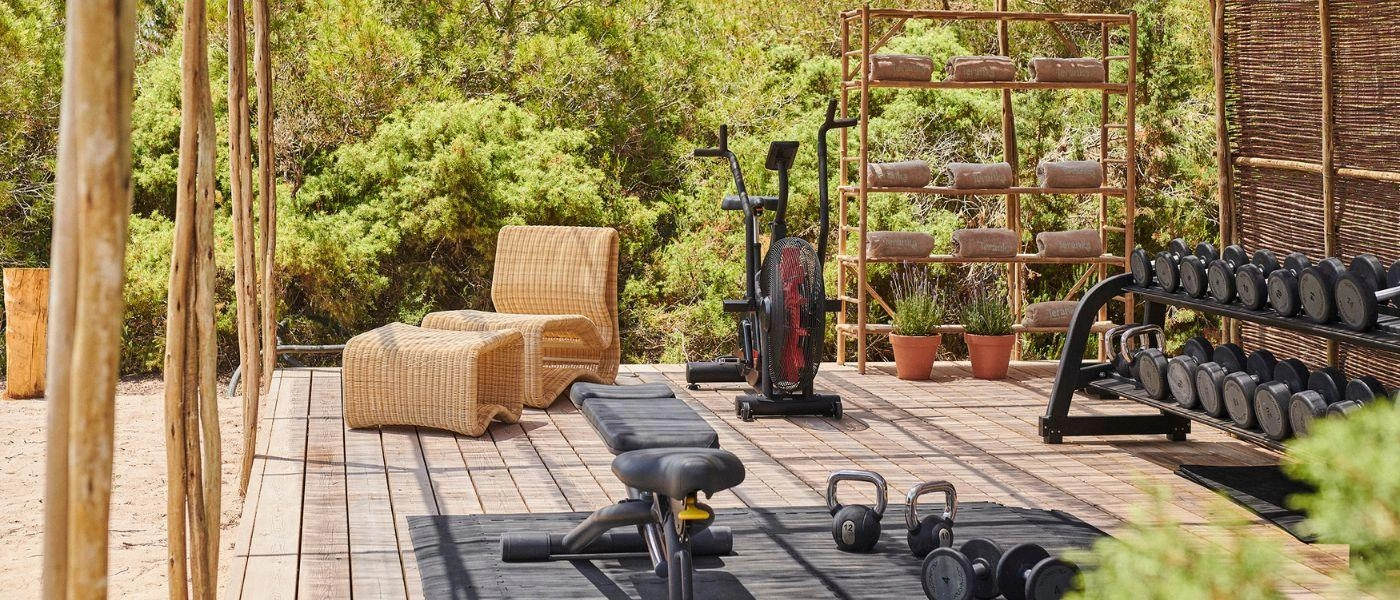 Open-air gym surrounded by greenery, with stacks of free weights, an exercise bike and bench