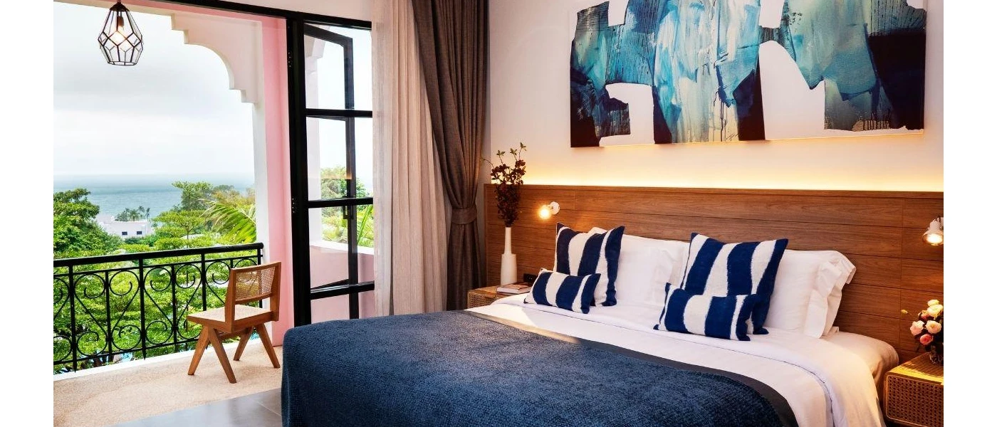 Hotel room with white bedding and walls, a navy throw and blue and white cushions, a wooden headboard and a blue and white wall painting, plus balcony doors open onto a balcony with greenery and sea views