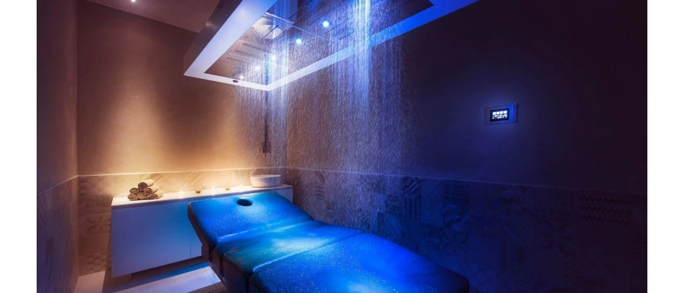 Treatment cabin with a cushioned bed, atmospheric blue lighting and a vanity unit with a sink and towels behind it