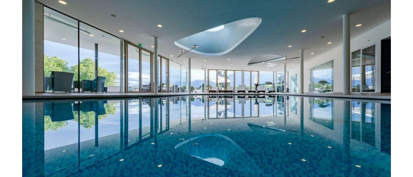 Large indoor pool surrounded by white-cushioned loungers under a white ceiling with spotlights, and floor-to-ceiling windows showing the greenery outside