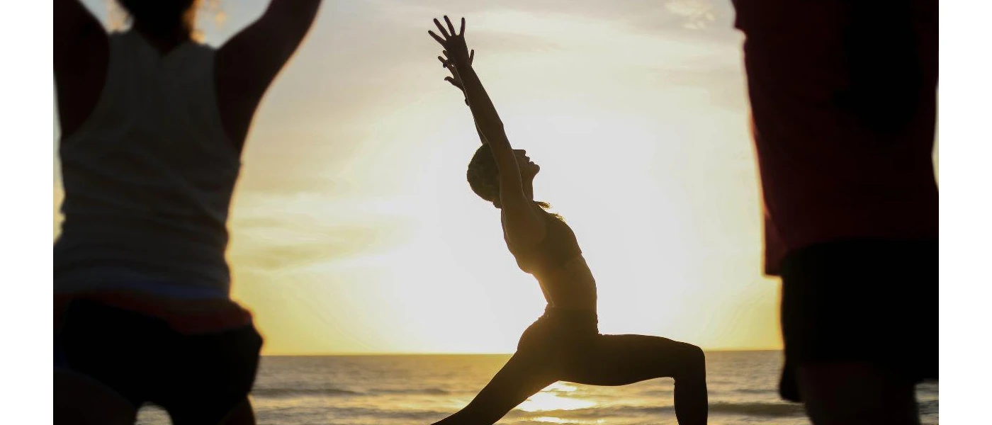 Silhouette of three people in yoga poses on the beach as the sun sits just above the ocean horizon