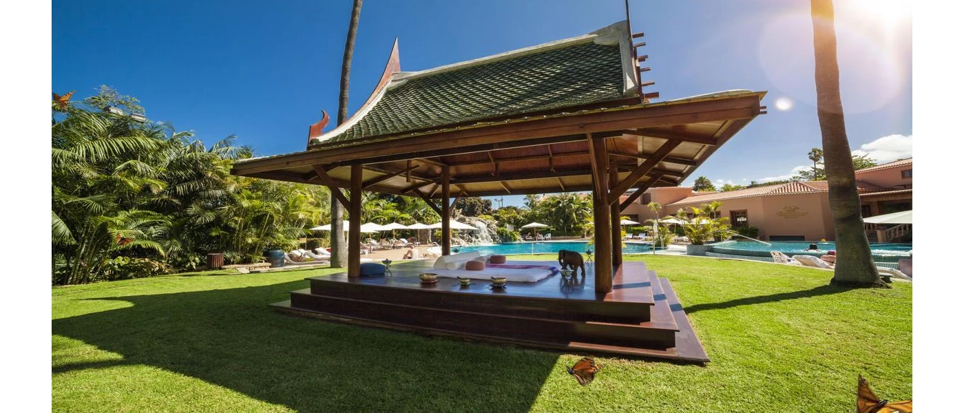 Wooden open-sided cabana with an Asia-style rooftop in a sunny garden with a lawn and swimming pools