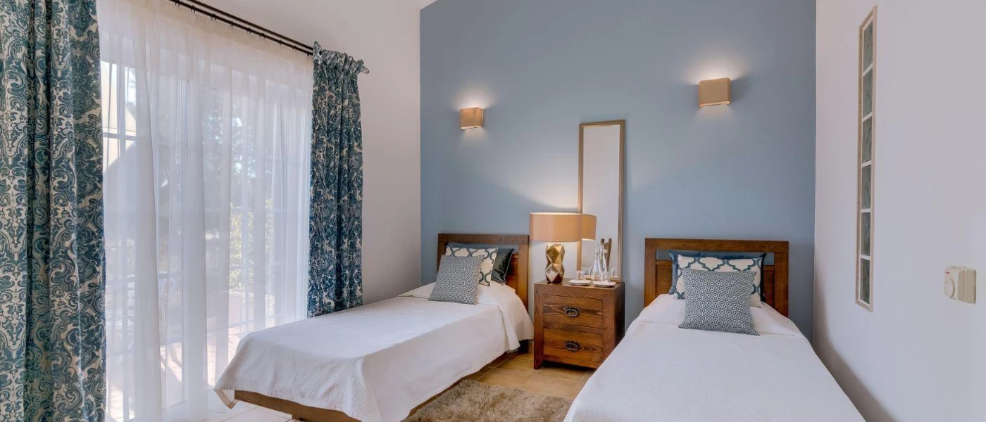 Twin bedroom with white bedding and blue cushions, a blue wall and blue and white sheer curtains