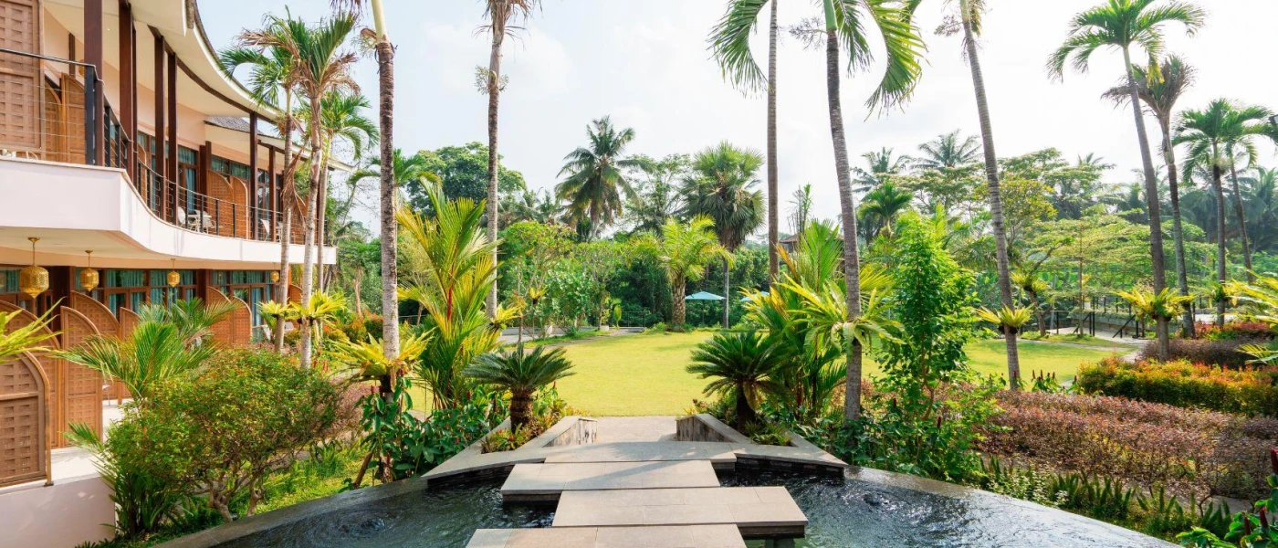 Paved slabs over water, leading to a garden filled with palms and overlooked by a rose-coloured building with rattan 