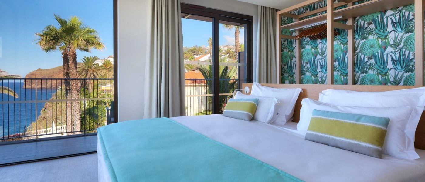 Modern hotel room with white bedding with a blue throw and striped cushions, tropical wallpaper and a view of the sea and cliffs