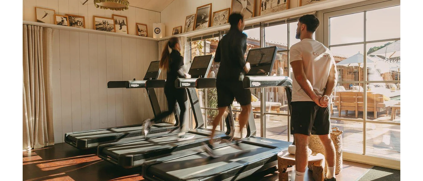 Man and woman in activewear run on treadmills in a bright studio, watched over by a fitness professional