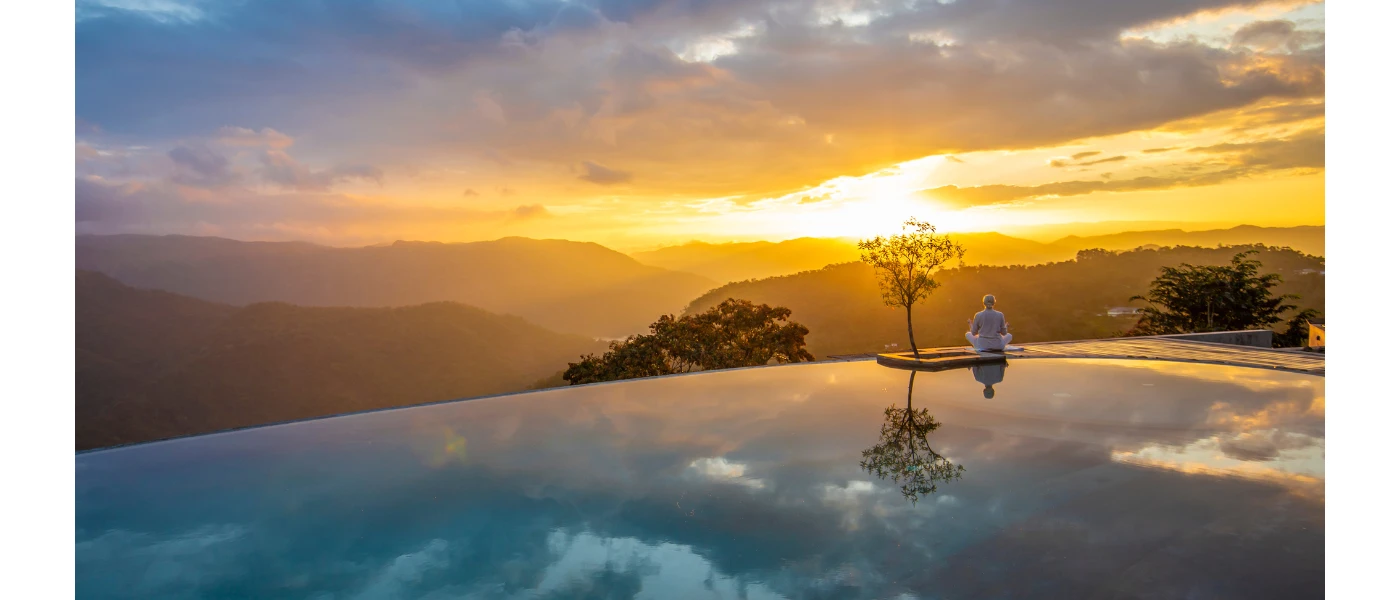 Infinity-edge pool at sunset, with mountains in the distance and a guest meditating on the edge