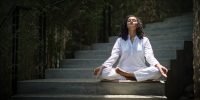 Woman in white sits cross legged with her eyes closed and palms up on a stone staircase