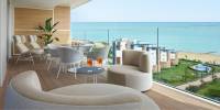 Glass-fronted balcony overlooking gardens and the sea, with neutral chairs and loungers and a table with aperol spritz