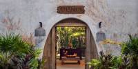 Spa at Palmaïa – The House of AïA Stone-walled entrance to the Atlantis Spa, with an arched entrance fringed by greenery peering through to a wooden table covered in candles