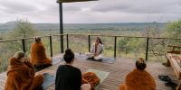 Group on yoga mats in brown blankets sit cross legged on a wooden deck overlooking the African wilderness