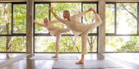 Man and woman in white practice yoga in an airy studio with wraparound windows and tropical greenery beyond