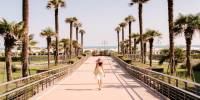 Woman in white dress and straw hat strolls across a paved bridge lined by palms towards the sea