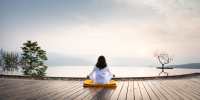 Woman sits on the edge of an infinity pool on a wooden deck meditating with misty hills in the distance