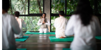 Group in white sit with their hands clasped in prayer on green yoga mats in a wooden studio