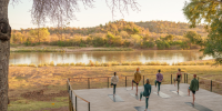 Group participate in a class on a wooden deck overlooking a river and thick greenery