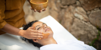 Woman lies on a bed in a white robe with her eyes closed as a therapist spreads an orange mask on her face