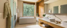 Neutral bathroom with wooden furniture and wall panels, a white standalone bathtub next to a large window, his and hers white sinks and a beige robe hanging on a hook, 