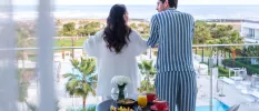 Dark-haired couple in pyjamas standing on a glass balcony looking down on grounds covered in palm trees and a central swimming pool