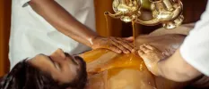 Man lies on his back as oil is poured onto his chest and two therapists guide the stream