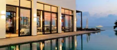 Twilight shot of white building with seating and large windows, overlooking the edge of a swimming pool and the ocean