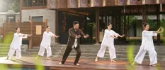 Four women in white follow an instructor in brown on a wooden poolside terrace
