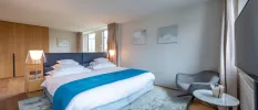 Modern hotel bedroom with crisp white bedding with a blue headboard and throw, a grey patterned rug underneath, light-wood wardrobe, a grey corner chair and table and a wall-hung TV