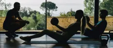 Male and female work out with medicine balls on mats in a studio with floor-to-ceiling windows, as a trainer watches from the front