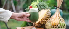 Glass of green juice with a cherry rested on top, next to a basket of fruit and a large pineapple