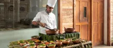 Chef in whites prepares food on an outdoor cooking station covered in bamboo leaves and wooden bowls