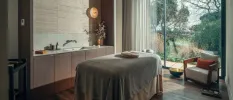 Stylish spa treatment room with wooden flooring and wood panelling, a floor-to-ceiling window, a bed with grey towels on it and a vanity unit with a vase of pink flowers on top
