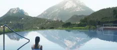 Dark-haired woman in a white swimsuit in an infinity pool overlook green and snowy peaks