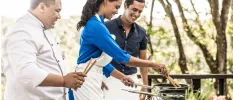 Man, woman and chef in whites smiling together as they cook with pans over a hob in the open air