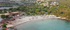 Milky beach furnished with loungers and parasols, surrounded by greenery and overlooked by white buildings