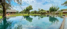 Rectangular swimming pool surrounded by lawns, trees and thatched-roof buildings under a blue sky