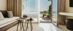 Hotel bedroom living area with a neutral sofa and cushions, round rattan rug, dark circular coffee tables and a wall-hung TV, plus a balcony with sea views