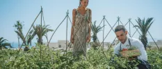 Woman in floral dress watches as a staff member picks herbs and vegetables from a kitchen garden under a blue sky
