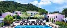 Swimming pool next to a paved terrace with white parasols and palm trees, surrounded by pink buildings