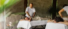 Man and woman lie on their fronts on massage beds in an open-air cabana surrounded by greenery, as two therapists massage their backs