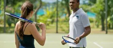 Woman with a tennis racquet standing next to a man with a tennis racquet on an ocean-facing court 