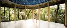 Woman in a white shirt dress in a yoga pose in the middle of an open-air pavilion surrounded by tropical greenery