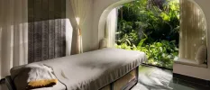 Spa treatment room with a massage bed draped in grey fabric, and a large curved window that looks out to tropical gardens