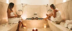 Two women chatting in a steam bath in white towels with candlelight and fluffy white spare towels