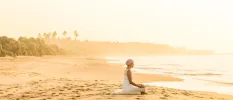 Woman in a white dress and headscarf sitting on a cushion on the beach, gazing out to sea as the sun rises 