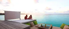 Couple sit on a cushioned terrace area, next to sofas with cream and green cushions overlooking their private pool and the ocean