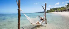 Woman in a white swimsuit and a straw hat lying in a hammock in clear shallows next to a sandy beach