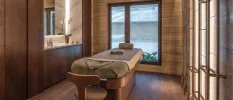 Wood-panelled spa treatment room with a massage bed and neighbouring sink and mirror, plus a large window behind